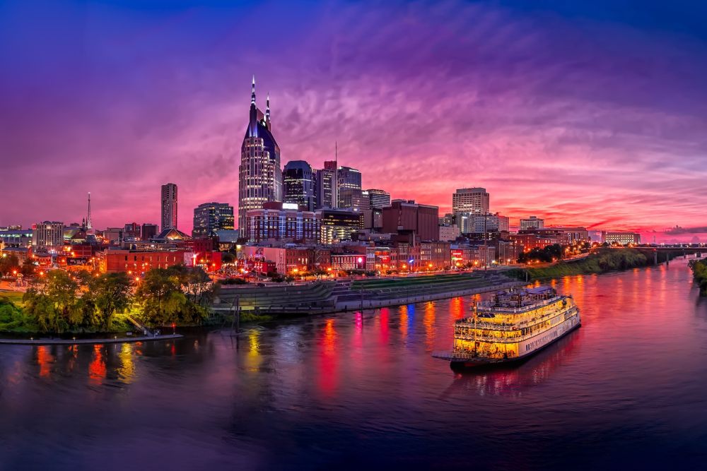 Nashville skyline at sunset with city lights reflecting on the river and a lit-up riverboat cruising by.